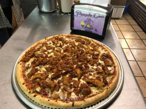 A large pizza topped with chunks of chicken on a metal tray, next to a sign for "Purple Cow."