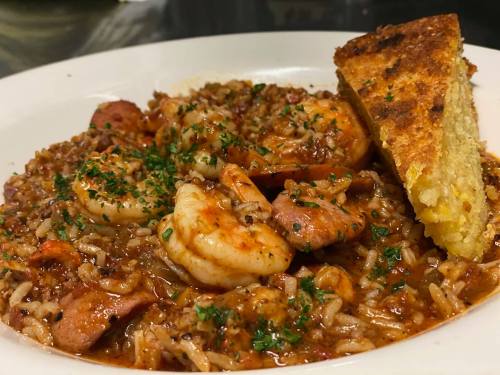 A plate of shrimp jambalaya with sausage, rice, and a slice of cornbread, garnished with parsley.