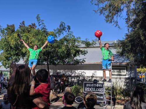 Two performers in bright green shirts juggle colorful balls while balancing on a ladder at an outdoor event.