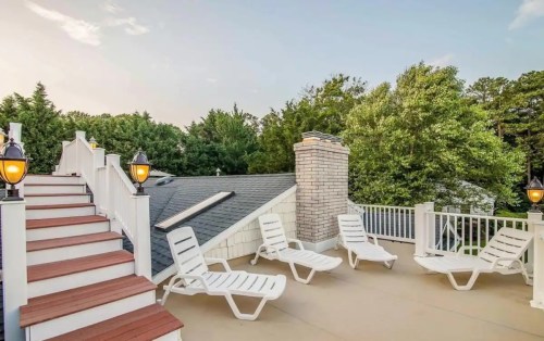Rooftop terrace with lounge chairs, stairs, and a chimney, surrounded by trees under a clear sky.