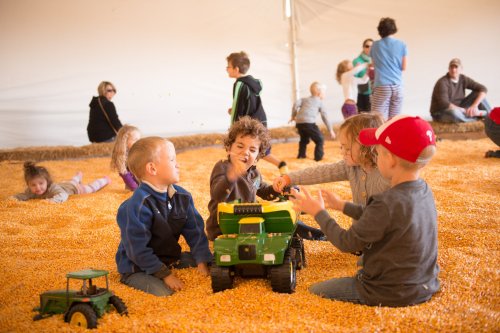 Children play with toy trucks in a corn pit, surrounded by others enjoying the activity in a large tent.