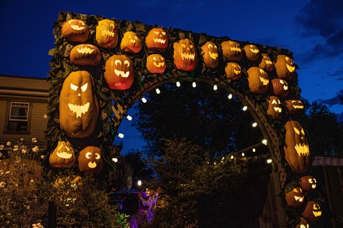 A spooky archway made of carved pumpkins with various faces, illuminated at night with string lights.