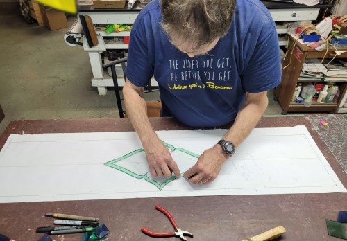 A person in a blue shirt sketches a design on a large piece of paper, surrounded by crafting tools.