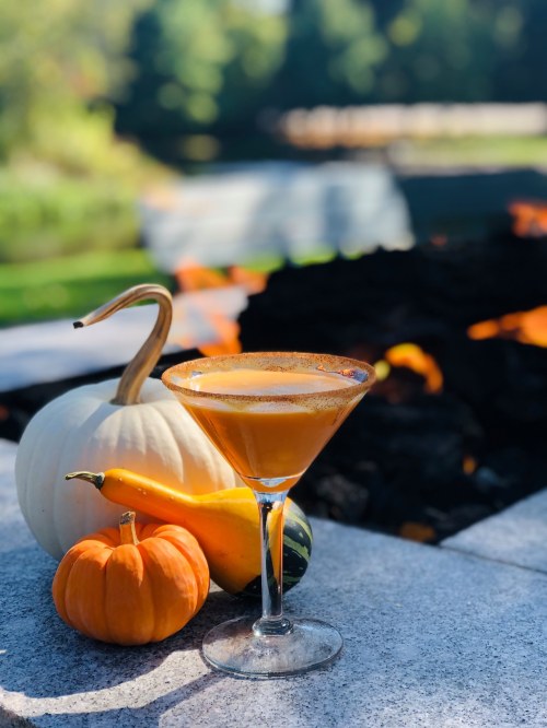 A pumpkin-themed cocktail sits in a glass near decorative pumpkins and a fire pit in a sunny outdoor setting.