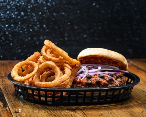 A basket of crispy onion rings next to a burger topped with onions on a wooden table.