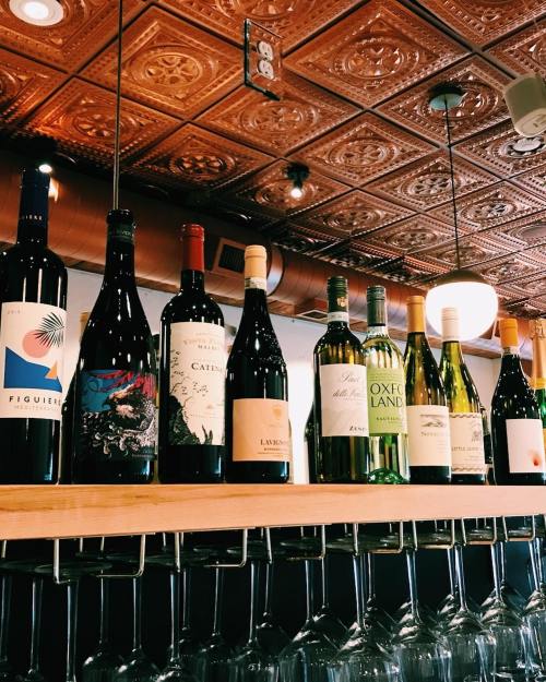 A row of various wine bottles displayed on a wooden shelf above a line of wine glasses in a cozy bar setting.