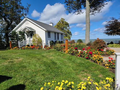 A charming white house surrounded by colorful flower beds and lush green grass under a blue sky with fluffy clouds.