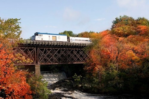 A train crosses a wooden bridge surrounded by vibrant autumn foliage and a flowing river below.
