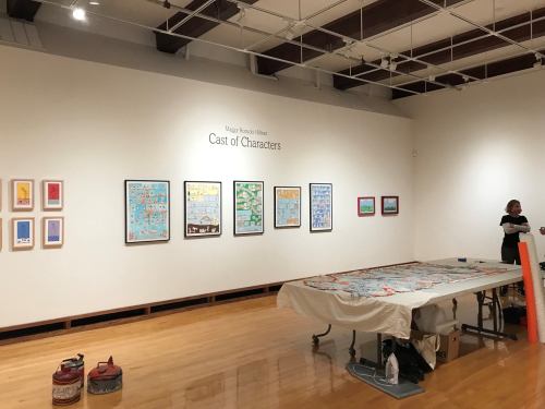 Art gallery interior featuring a wall of framed artworks titled "Cast of Characters" and a table with art supplies.