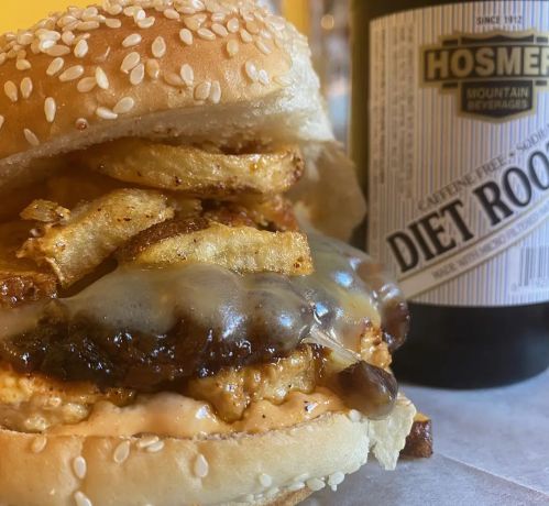 A close-up of a sesame seed burger with melted cheese, crispy fries, and a bottle of diet root beer in the background.