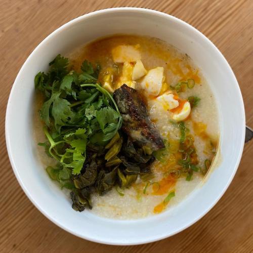 A bowl of congee topped with greens, cilantro, egg, and pieces of fish, served on a wooden surface.