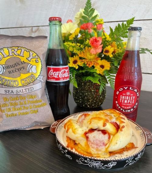 A table setting with a bag of potato chips, two soda bottles, and a plate of food, alongside a colorful flower bouquet.