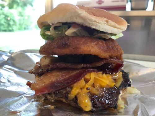 A towering burger with layers of bacon, cheese, fried chicken, lettuce, and a soft bun, all wrapped in foil.