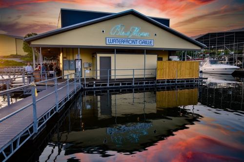 A waterfront restaurant with a neon sign, reflecting in the water at sunset, surrounded by boats and a colorful sky.