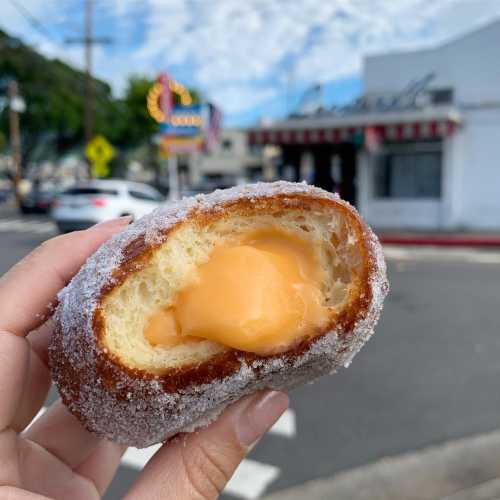 A hand holds a sugar-coated donut filled with orange cream, with a blurred background of a street and buildings.