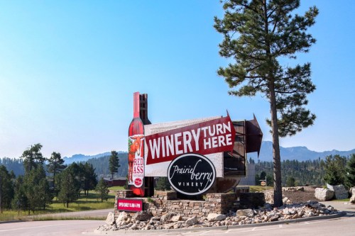 Sign for Prairie Berry Winery with a large wine bottle graphic, surrounded by trees and mountains in the background.