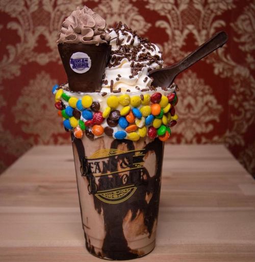 A tall, decadent milkshake topped with whipped cream, chocolate sprinkles, and a mini dessert cup, surrounded by colorful candies.
