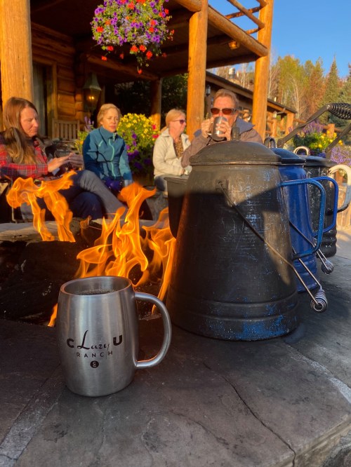 A cozy outdoor scene with people sitting around a fire, a kettle, and a mug in front of a rustic log cabin.