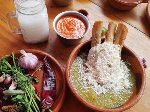 A bowl of green soup topped with cheese and breadsticks, accompanied by a side of salsa, herbs, and a glass of drink.
