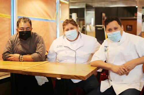 Three chefs wearing masks sit at a wooden table in a restaurant, smiling at the camera.