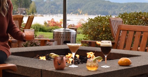 A cozy outdoor setting with a fire pit, featuring various cocktails and a scenic lake view in the background.
