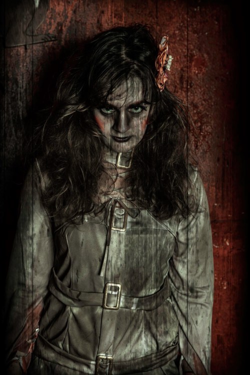 A woman in a straitjacket with disheveled hair and eerie makeup stands against a dark, textured background.