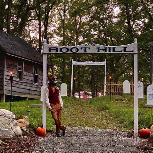 A person in a vintage outfit stands near a sign reading "Boot Hill," surrounded by pumpkins and a spooky graveyard.