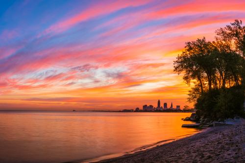Sunset over a calm lake with vibrant pink and orange skies, silhouetting a city skyline and trees along the shore.
