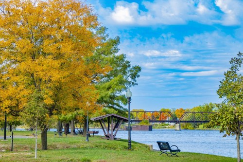 A scenic riverside park with colorful autumn trees, a bench, and a bridge in the background under a blue sky.