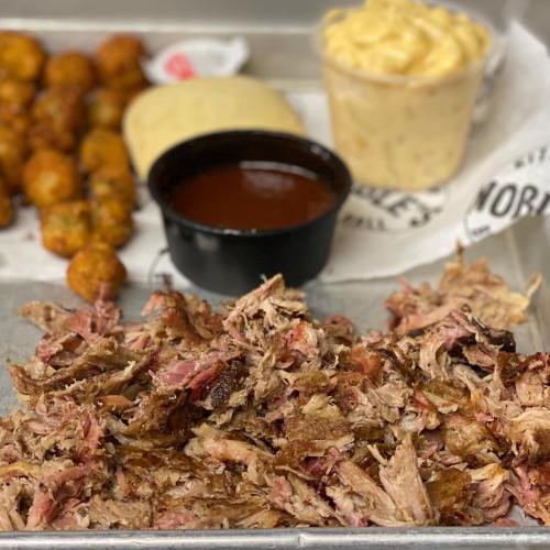 A platter featuring pulled pork, a side of barbecue sauce, hushpuppies, cornbread, and macaroni and cheese.