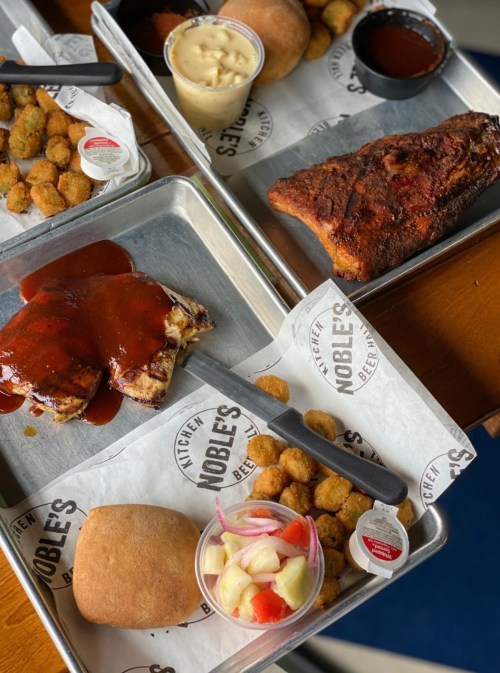 A spread of barbecue dishes on metal trays, featuring ribs, sides, rolls, and dipping sauces.