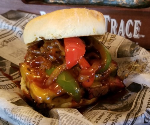 A hearty burger topped with grilled peppers and onions, served on a bun with barbecue sauce, wrapped in newspaper.