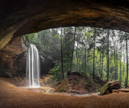 A serene waterfall cascades from a rocky overhang into a lush, green forest setting.