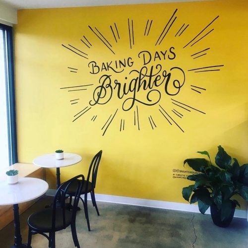 A bright yellow wall with the text "Baking Days Brighter" and two small tables with chairs in a cozy space.