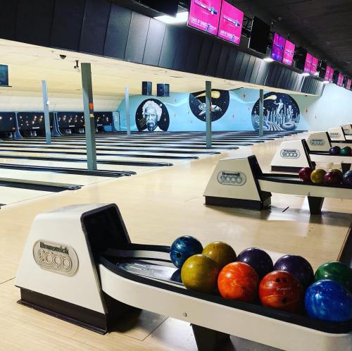 A bowling alley with colorful balls on a rack and screens displaying scores in the background.