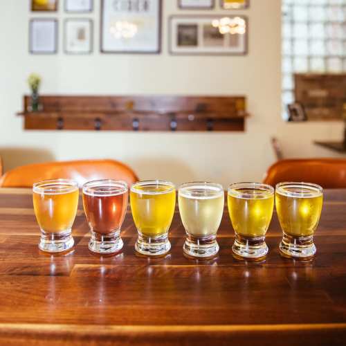 A row of six small glasses filled with different colored beverages on a wooden table, with a cozy background.