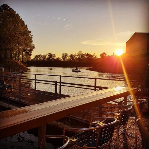 Sunset over a calm waterway, with boats passing by and a wooden deck lined with chairs.