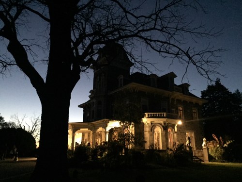 A dark silhouette of a large, historic house illuminated at night, surrounded by trees and a twilight sky.
