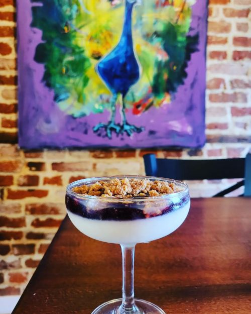 A dessert in a glass with a crumb topping, set against a colorful peacock painting on a brick wall.
