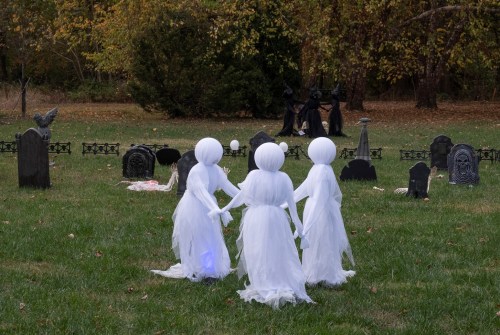 Three ghostly figures in white costumes hold hands in a graveyard, surrounded by tombstones and autumn foliage.