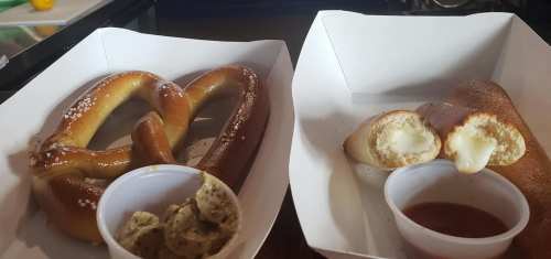 Two pretzels on white trays: one soft pretzel with salt and a stuffed pretzel with cheese, served with dipping sauces.