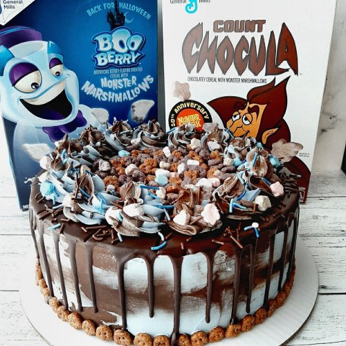 A chocolate cake decorated with blue and brown frosting, surrounded by cereal pieces, next to Boo Berry and Count Chocula cereal boxes.