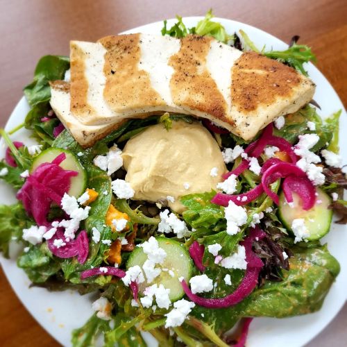 A colorful salad topped with grilled chicken, hummus, cucumbers, pickled onions, and crumbled feta cheese.