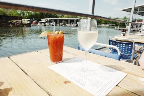 A wooden table by the water with a Bloody Mary and a glass of white wine, alongside a menu.