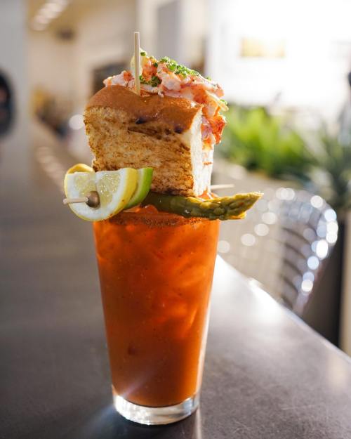 A tall glass of Bloody Mary topped with a slice of bread, lobster, lemon, lime, and pickles on a bar counter.