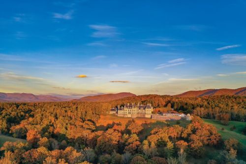 Aerial view of a grand mansion surrounded by vibrant autumn foliage and rolling hills under a clear blue sky.