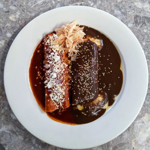 Two enchiladas on a plate, one topped with cheese and onions, served with a rich dark sauce and sesame seeds.