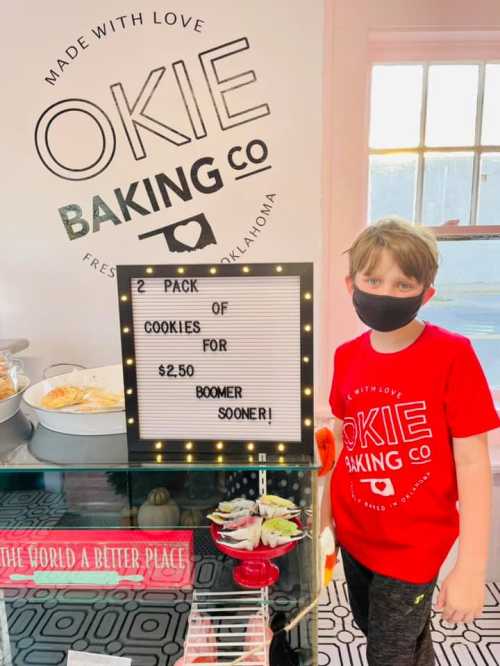 A child in a red shirt stands next to a sign advertising a two-pack of cookies for $2.50 at Okie Baking Co.