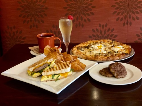 A plate with a sandwich, chips, a cocktail, a pizza, and a plate of sausage patties on a wooden table.
