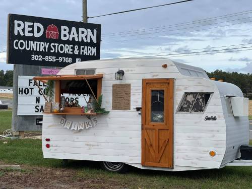 A vintage white camper trailer with a wooden window serving drinks, parked near a sign for Red Barn Country Store & Farm.